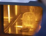  Close-up of a 3D printing process inside a machine, showing a partially formed anatomical model illuminated by warm orange lighting within an enclosed chamber.