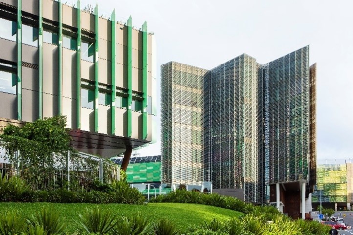 Queensland Children's Hospital buildings with distinctive architectural designs, including one structure with vertical green shading fins and another with a patterned, perforated façade. In the foreground, landscaped greenery with shrubs and grass slopes leads up to the buildings. A roadway with a few cars is partially visible to the right under a lightly overcast sky.