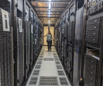 A narrow aisle inside a server data centre with tall racks of black computing equipment and network servers on both sides, connected by visible cabling. A person stands at the far end of the corridor, facing towards the servers. The floor features a grated metal walkway, and overhead lighting evenly illuminates the tightly arranged infrastructure.