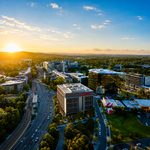 The sun sets behind a city, illuminating buildings and trees with vibrant colors in the evening sky.