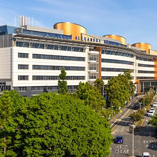 Princess Alexandra Hospital in Brisbane, a large modern multi-storey building with a curved façade and gold cylindrical features on the roof. The hospital name is visible in blue lettering along the верх of the building. In the foreground, dense green trees line a street with cars parked and driving along a clear roadway under a blue sky.