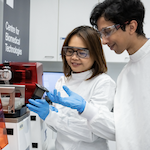 Two laboratory workers in white coats and safety glasses examine a small piece of equipment while wearing blue gloves beside a red 3D printer or lab device.