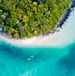 erial view of a tropical shoreline with lush green forest meeting a curved white sandy beach and clear turquoise water, with two small boats offshore.