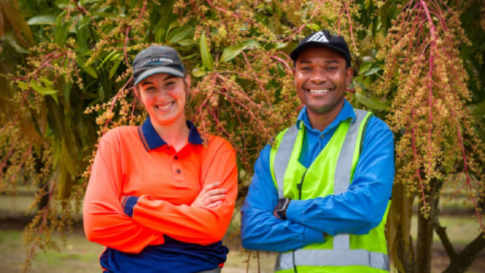 Two outdoor workers standing side by side with arms crossed, wearing high-visibility and work shirts, in front of leafy trees with hanging branches.