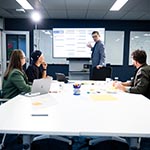 Photo of a person at a whiteboard talking with three seated people.